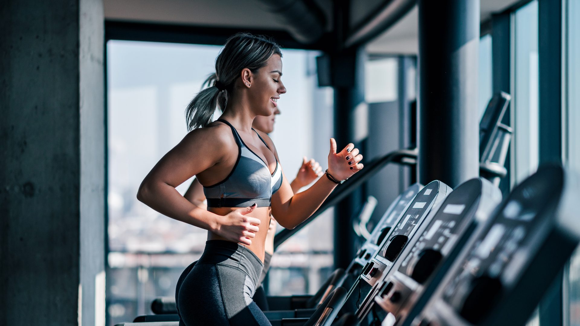 A side view of a women is a grey and black sports bra and shorts running on a treadmill burning calories