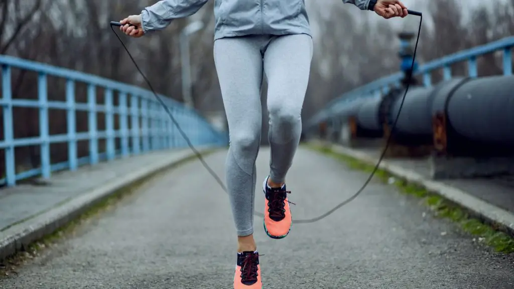 A woman outside on a bridge wearing grey exercise clothes and orange running shoes skipping rope 