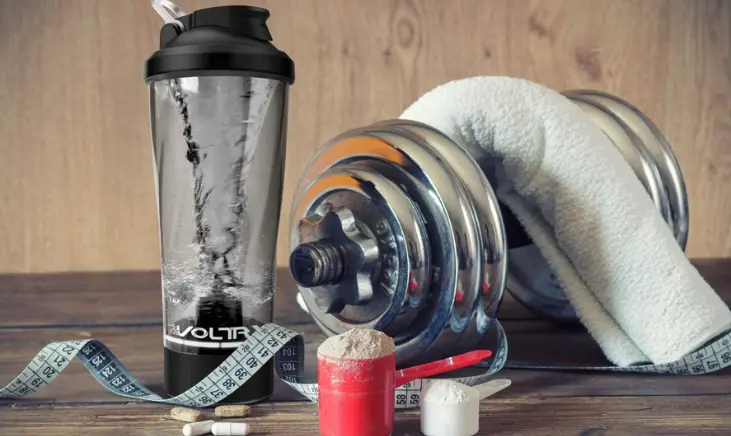 A black VOLTRX electric shaker bottle next to some supplements and an adjustable dumbbell with a towel resting overtop of the grip