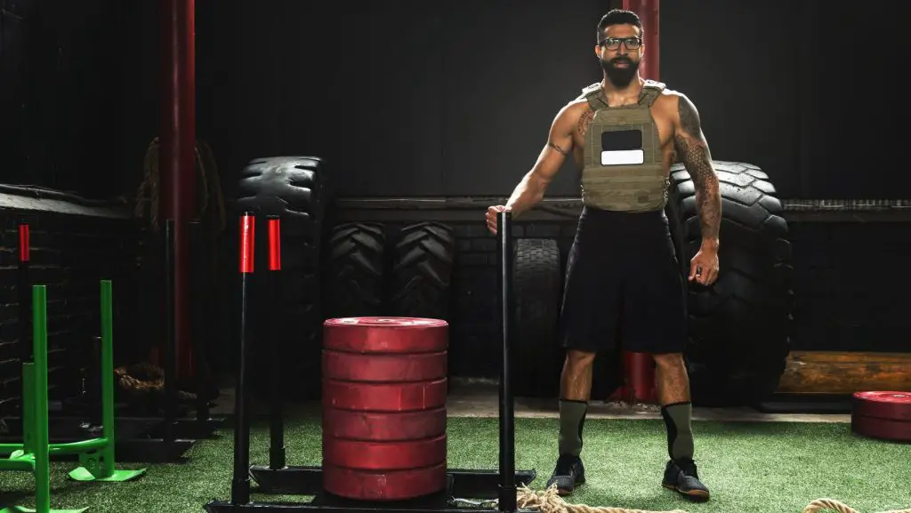 A man in a gym standing on turf, wearing a weighted vest while resting his hand on a gym sled 