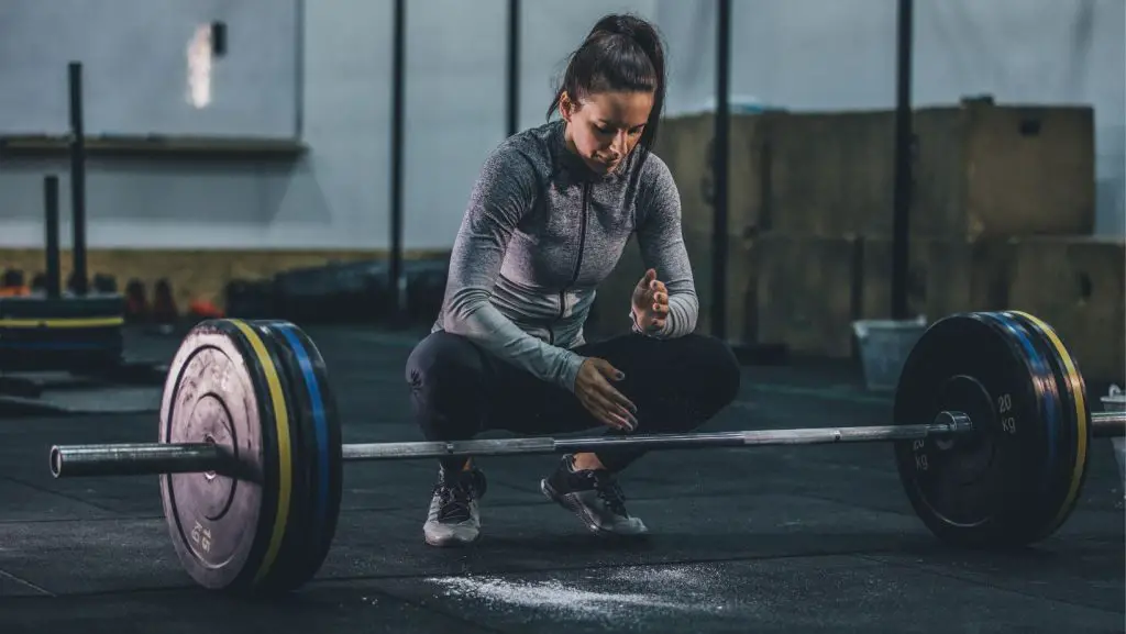 A woman squatting down behind a bar with plates in the gym, chalking her hands for grip in preparation for a deadlift  