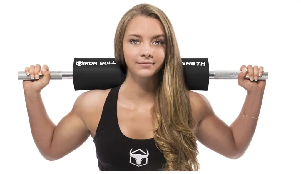 A woman facing forward, with a barball resting on her shoulders showcasing a black, Iron Bull Strength Advanced Squat Pad