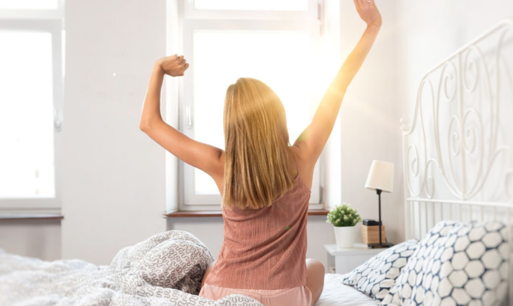 A woman stretching her arms after she has just woken up, the sun shining through her bedroom window