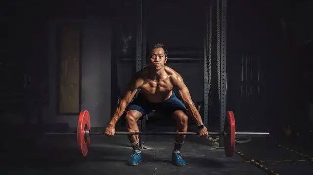 The photo shows a man standing in front of a barbell with his feet shoulder-width apart. His large, muscular quads are visible as he bends over to touch his shins to the barbell. 