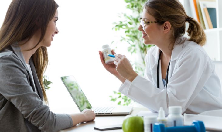 A photo of two women, one of whom is a doctor. The doctor is holding a tub of creatine gummies and is explaining something to the other woman.
