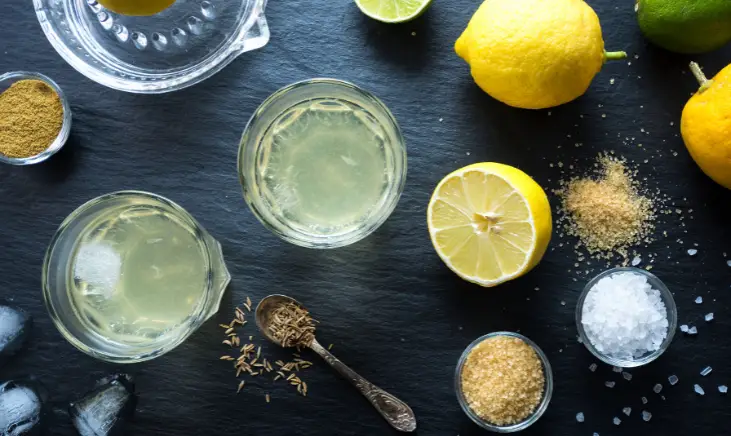 A glass of lemon juice being made with a lemon, a spoon, sugar, and seeds.