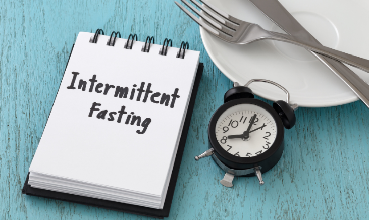 A notebook with the words "intermittent fasting" written on it, next to an alarm clock and a fork. 